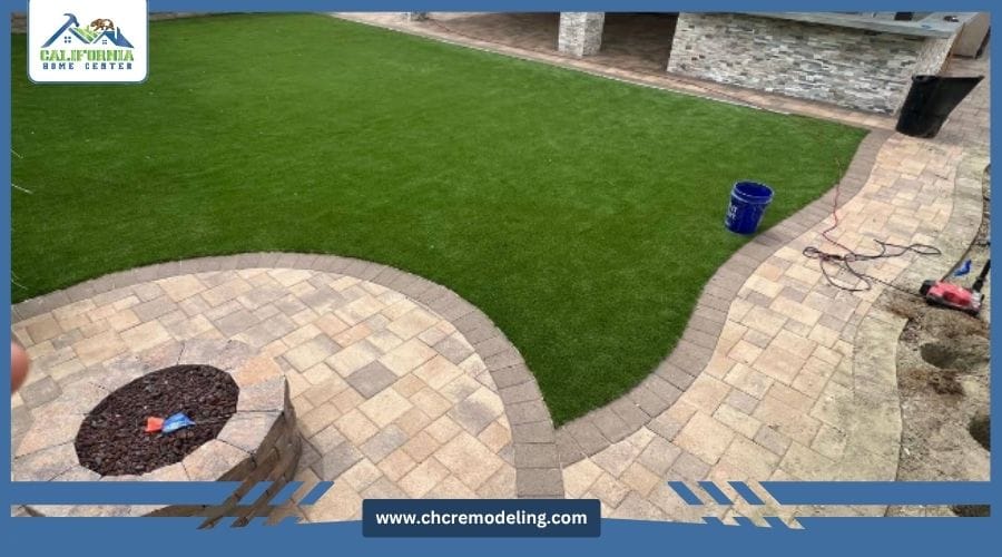 A landscaped backyard featuring a curved stone paver patio with a built-in fire pit next to a lush green lawn.